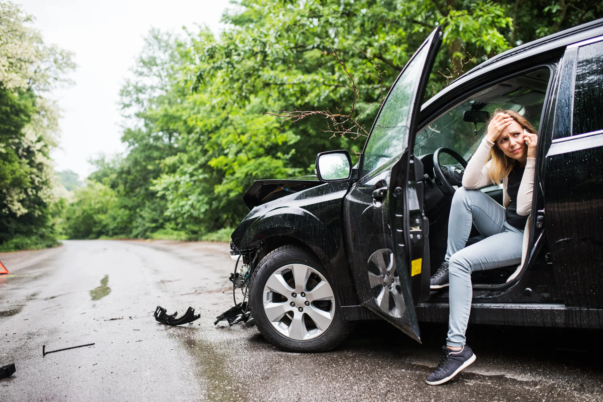 Woman sitting beside a damaged car on a wet road after a collision